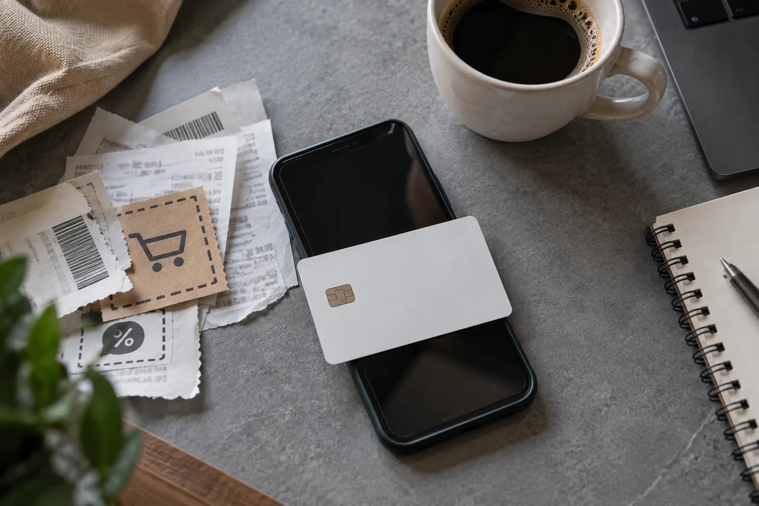 A smartphone, a blank debit-style card, a torn paper slip and a coffee laid untidily on a mid-grey table