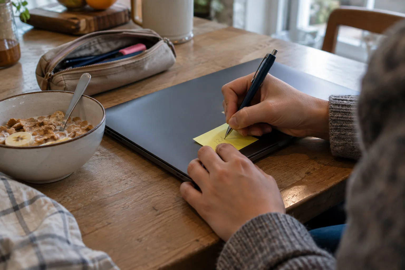Hands writing a number on a yellow sticky note stuck to a closed laptop on a kitchen table